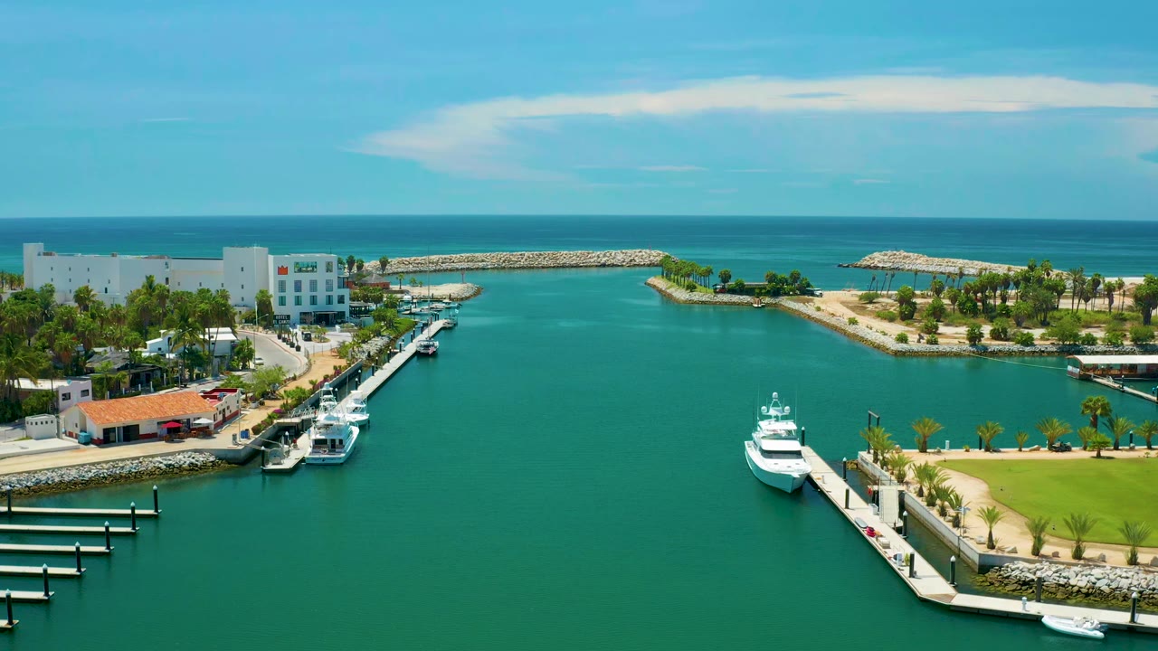 Opening aerial frame over San Jose del Cabo with teal marina water in front, white yachts at docks, low waterfront buildings, palm greenery, breakwaters, ocean horizon, and no visible people.