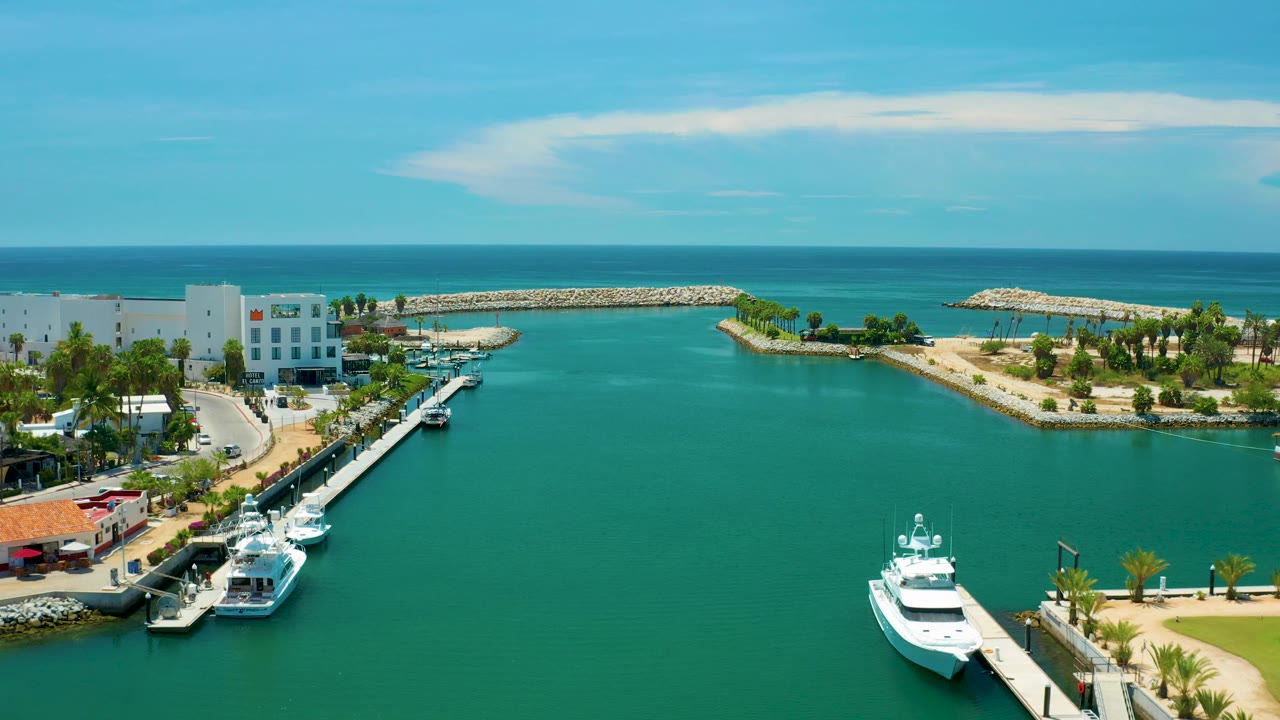 High view over San Jose Del Cabo harbor water, with yachts near the lower right, a white building and palm-lined road at left, breakwaters ahead, and no visible people.