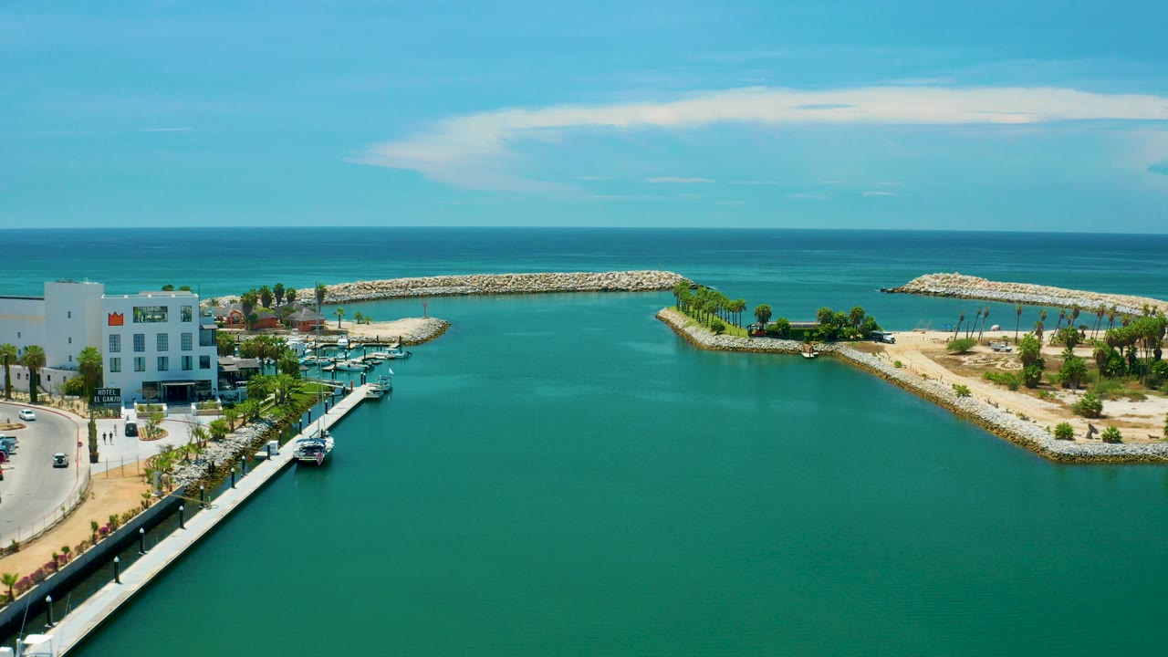High aerial frame over San Jose Del Cabo, Mexico, showing turquoise water, a marina beside white buildings, sandy breakwaters with palms, open ocean, blue sky, and no visible people.