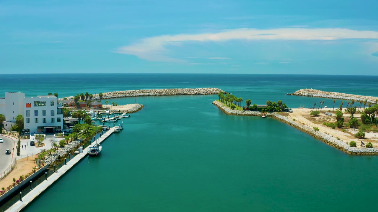 Wide aerial frame from San Jose Del Cabo, Mexico, showing a broad turquoise inlet, palm-lined sandbars and breakwaters, marina boats near white buildings, a flat ocean horizon, and no visible people.