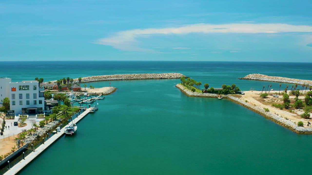 Aerial resort coastline with turquoise water, marina, white building edge, sandy breakwaters, palms, and blue ocean.