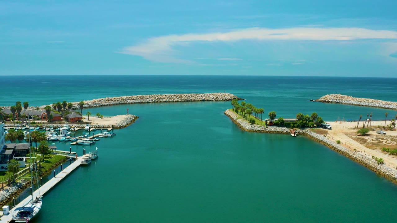 Aerial view in San Jose del Cabo, Mexico showing emerald lagoon water, marina docks with white yachts at lower left, a palm-lined point, stone breakwaters, blue ocean, and no visible people.