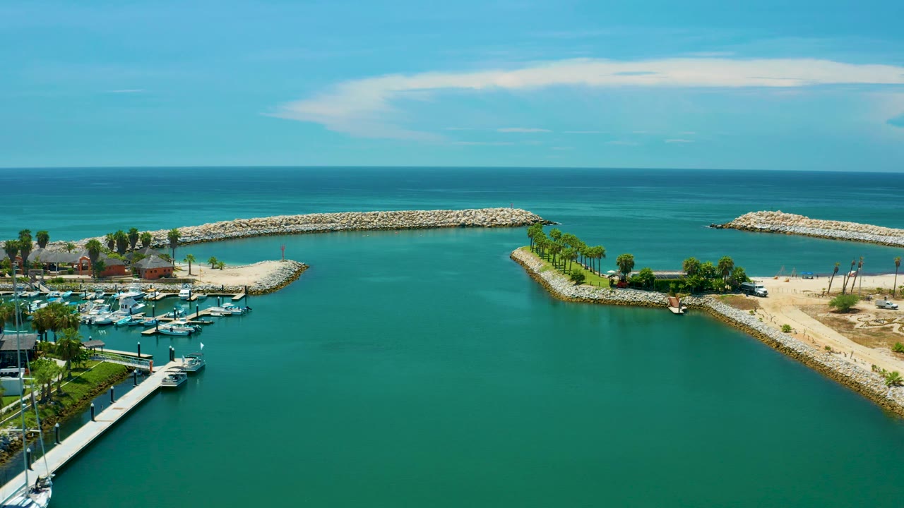 Aerial frame in San Jose del Cabo, Mexico showing the palm-lined marina point and breakwater across the center, with yacht slips at lower left, open turquoise water, ocean horizon, and no visible people.