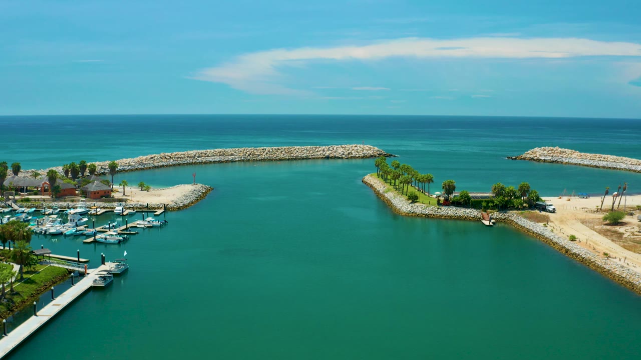 Aerial view in San Jose del Cabo, Mexico with a large expanse of green lagoon in the foreground, clustered yachts at lower left, a sandy point and stone breakwater at midframe, ocean beyond, and no visible people.