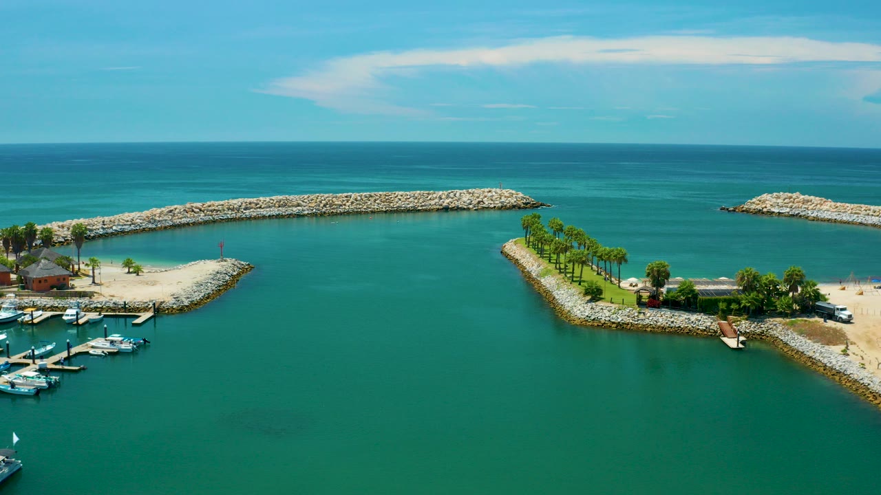 Aerial frame over San Jose Del Cabo showing turquoise water, pale rock breakwaters, a small marina dock area at lower left, coastal trees, blue sky, and no visible people.