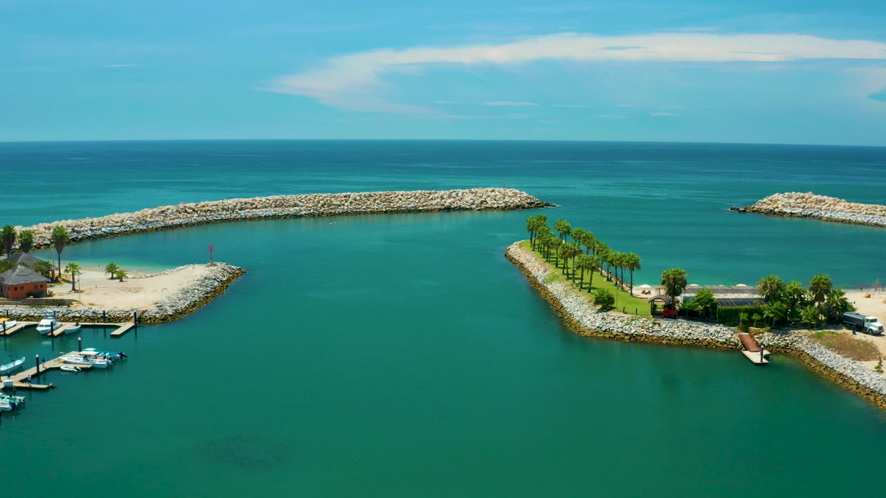 Aerial view over San Jose Del Cabo with open turquoise water, a long pale rock breakwater across the midground, a narrow tree-lined shore point at right, and no visible people.