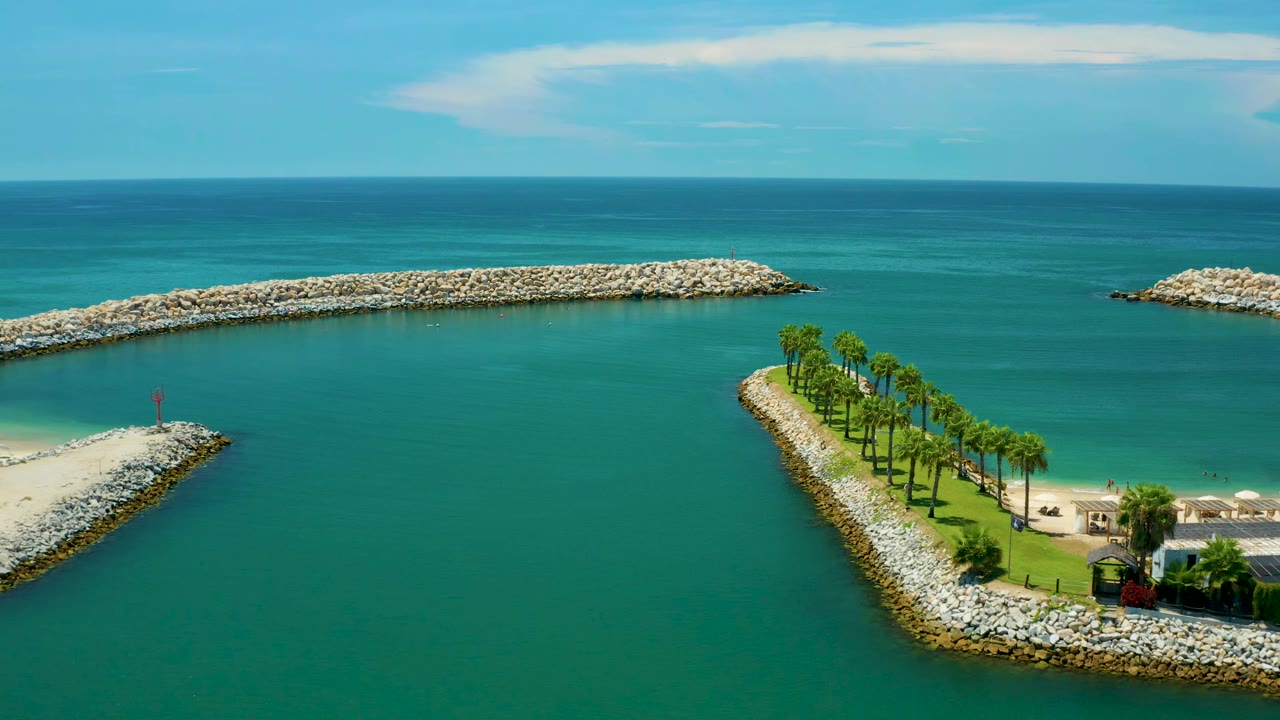 Aerial frame in San Jose Del Cabo, Mexico with the palm-lined rocky strip set lower in frame, open turquoise water in the center, two stone breakwaters near the horizon, and no visible people.