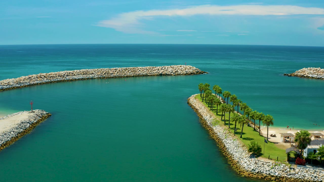 Aerial view in San Jose Del Cabo, Mexico showing the curl-shaped palm strip on the right, a small rocky point on the left, long offshore breakwaters, bright blue-green water, and no visible people.
