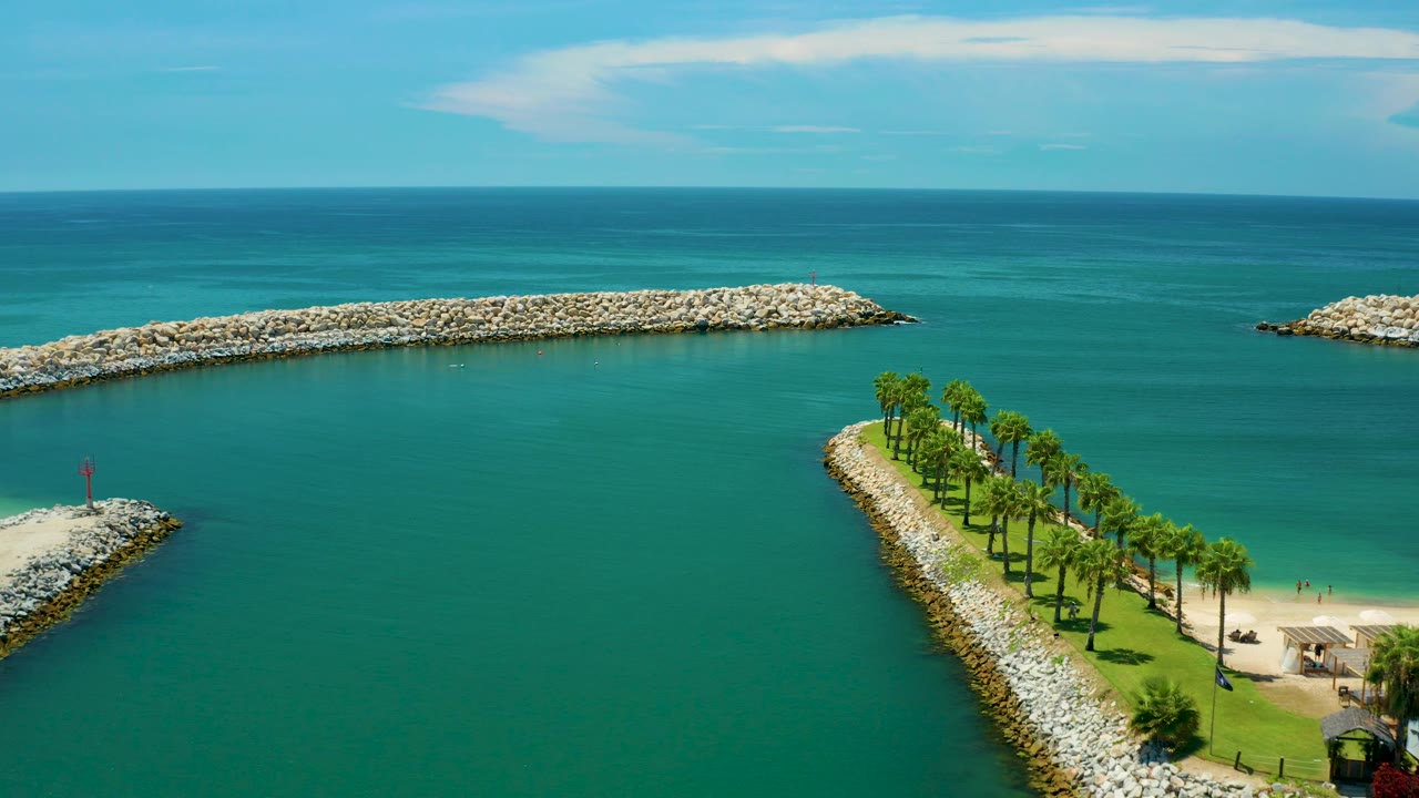 Wide aerial frame in San Jose Del Cabo, Mexico with a palm-covered rocky shoreline corner at lower right, a small rock jetty at lower left, open teal water, distant breakwaters, and no visible people.