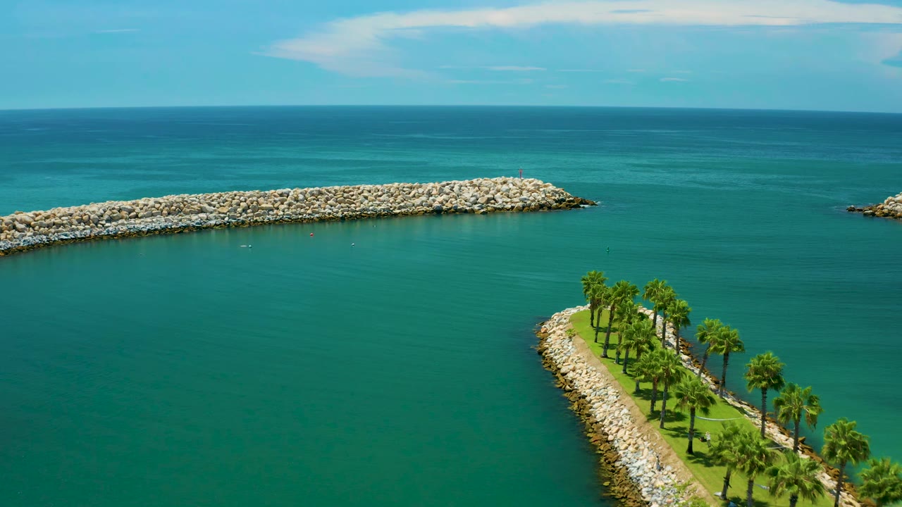 San Jose Del Cabo aerial view with the palm-covered point lower in frame, the rock breakwater centered across green-blue water, a distant horizon, and no visible people.