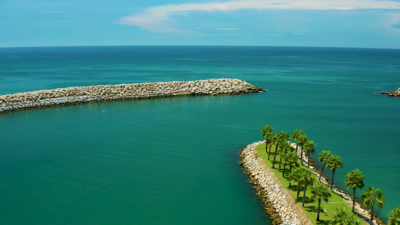 Aerial frame over San Jose Del Cabo with calm teal water around a curved palm-lined strip, a pale rock breakwater in the midground, open sea beyond, and no visible people.