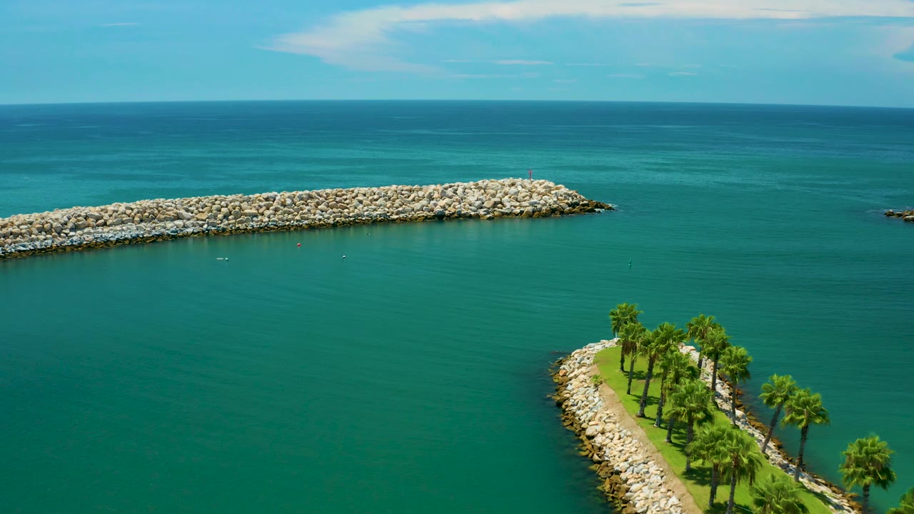 Mid-clip aerial view in San Jose Del Cabo with more open water visible, the rock breakwater stretching left to right, palms near the lower edge, and no visible people.