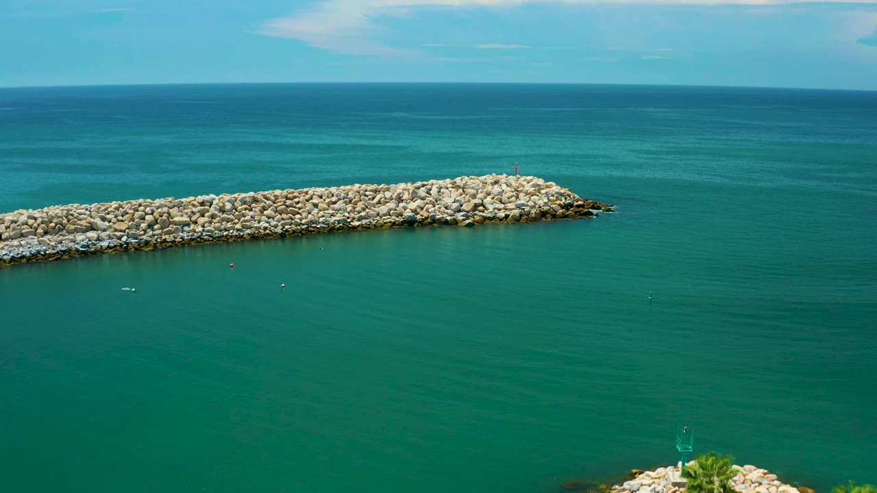Aerial-looking frame of a long rock jetty over green-blue ocean in San Jose Del Cabo, Mexico, with small floating markers and no visible people.