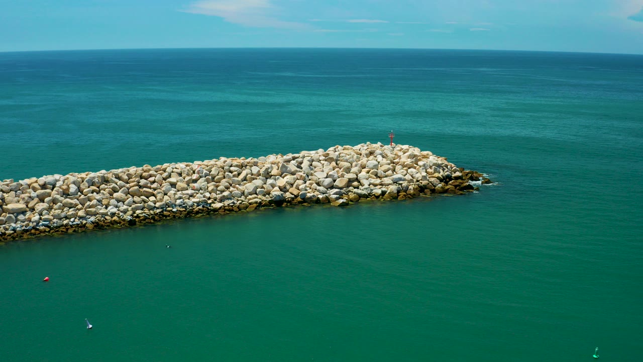 Aerial view in San Jose Del Cabo, Mexico, looking across a pale rock breakwater with teal ocean water, a high horizon, and a tiny distant water user near the lower left.
