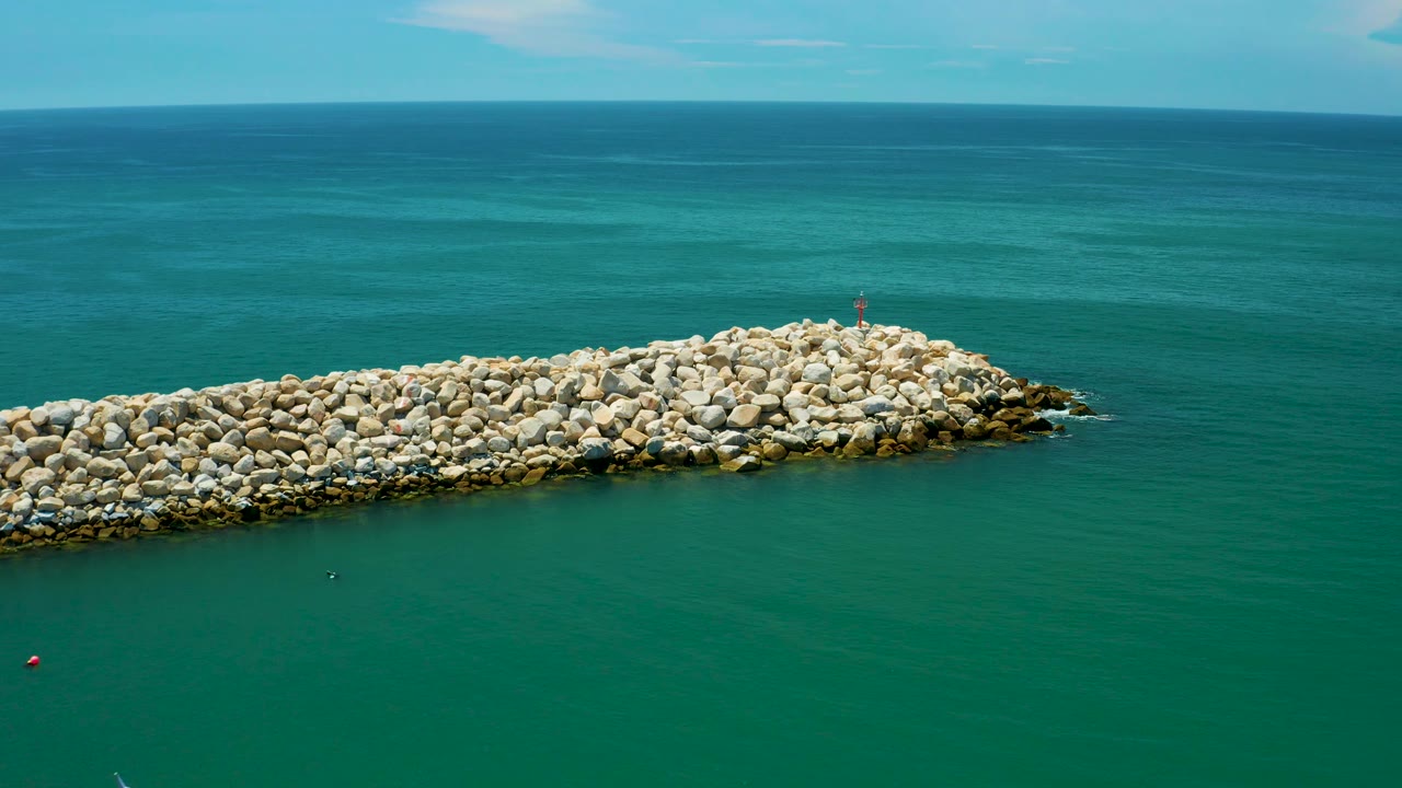 Aerial view in San Jose Del Cabo, Mexico, showing the boulder breakwater slightly farther right in frame, broad teal water, a level ocean horizon, and a small red marker near the tip.