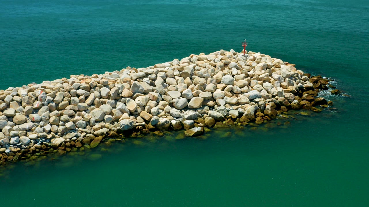 Aerial frame of a pale stone breakwater extending across blue-green water in San Jose del Cabo, Mexico, with a small red marker near the top edge and no visible people.
