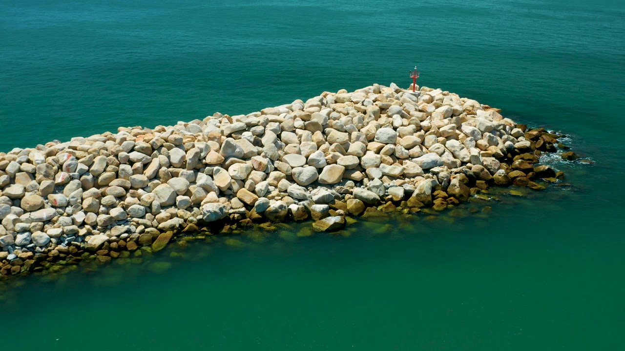 Aerial view in San Jose del Cabo, Mexico, with the stone dam centered lower in frame, turquoise water above it, darker rocks at the right edge, and no visible people.