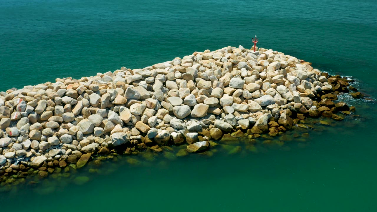 Aerial frame of the San Jose del Cabo stone breakwater pushed slightly right in composition, with open teal water behind it and no visible people.