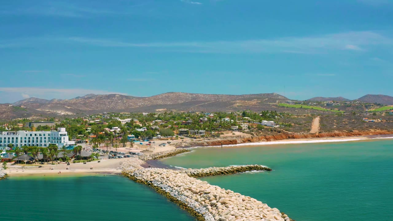 Aerial view of San Jose Del Cabo, Mexico with a rock jetty reaching into turquoise water, pale beach, white beachfront buildings, inland trees, brown hills, and no visible people.