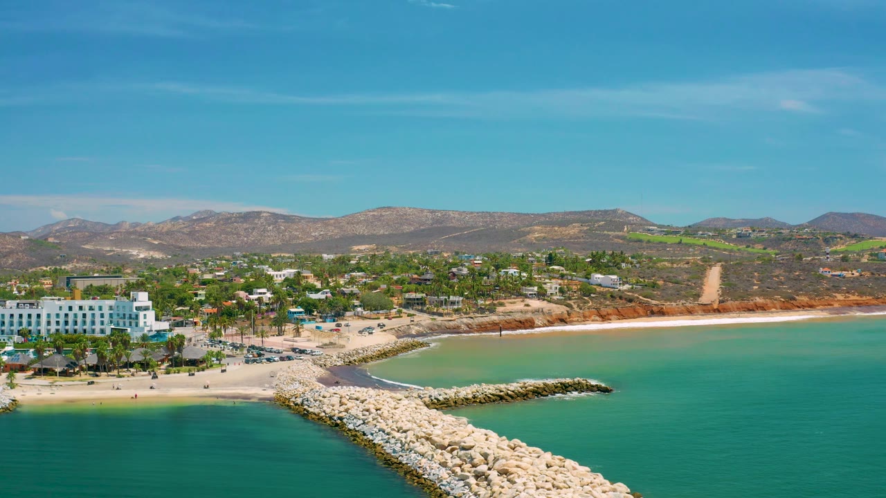 Drone view of San Jose Del Cabo, Mexico with a pale rock breakwater below, teal water, sandy shoreline, scattered town buildings, green trees, low brown hills, and no visible people.