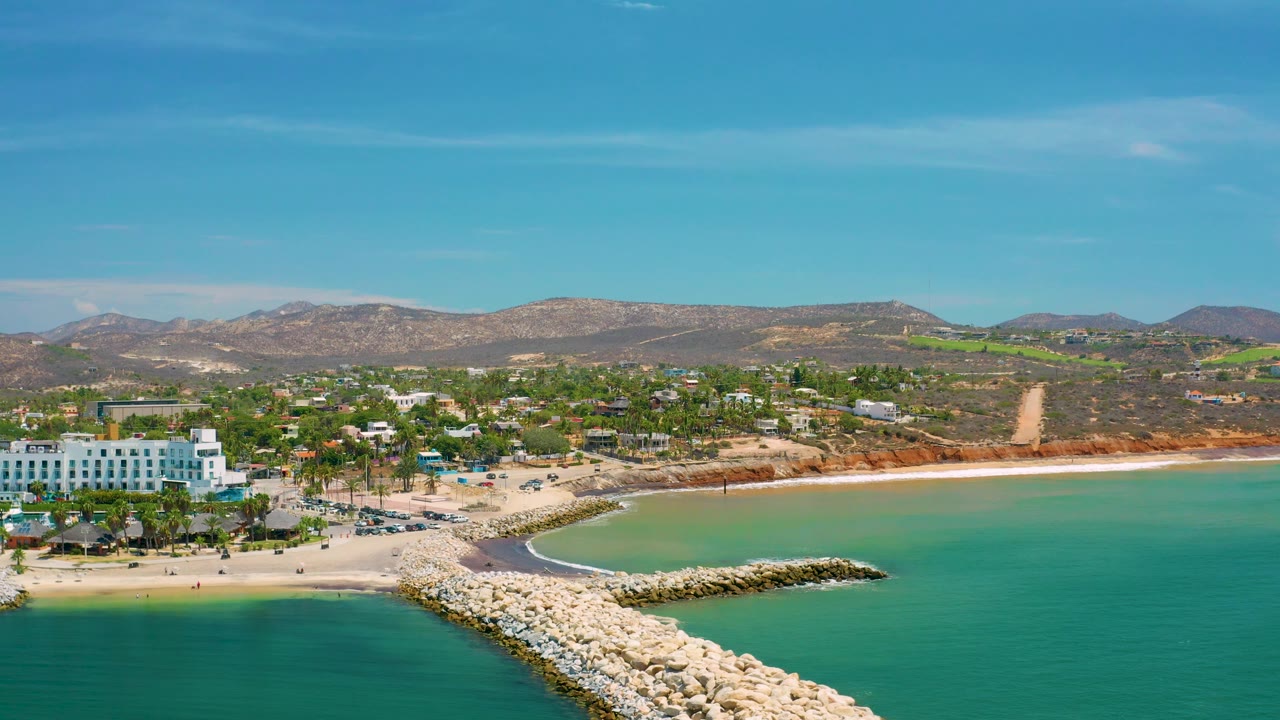 Aerial beach scene with white waterfront buildings, jetty rocks, turquoise water, and hills.
