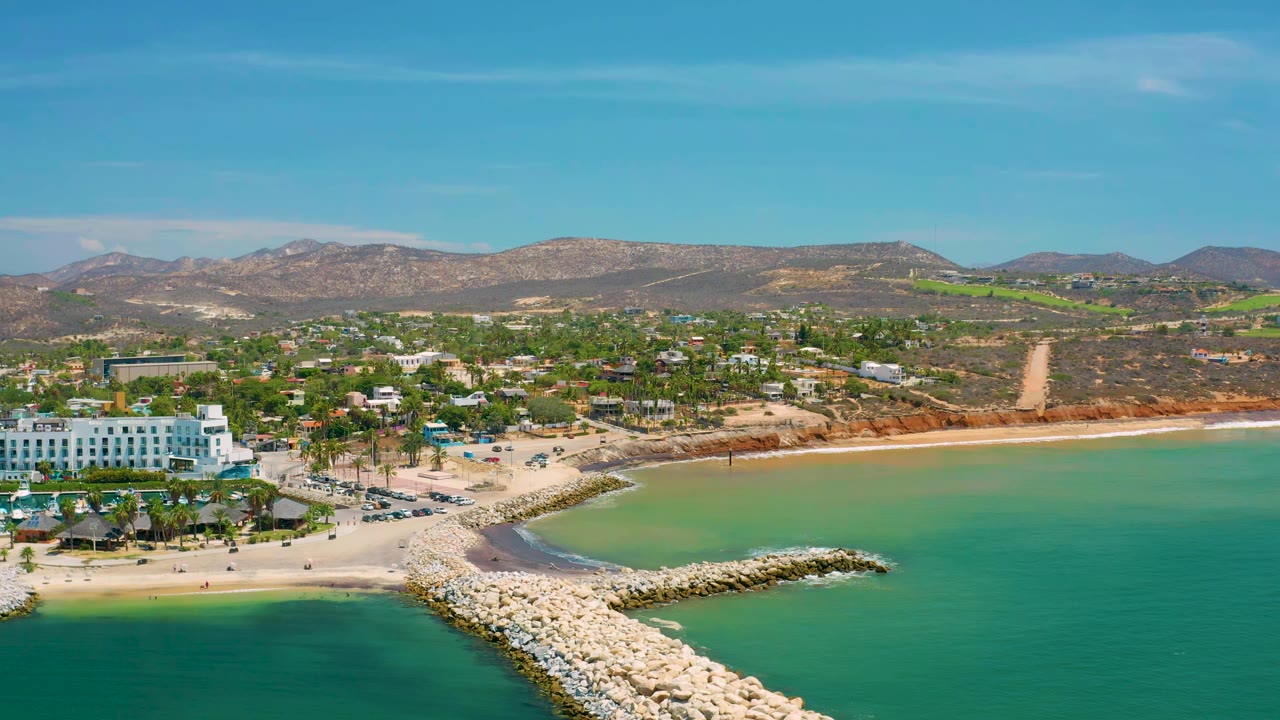 A second aerial view of San Jose Del Cabo frames the stone jetty lower center, shallow green water along the beach, white resort buildings to the left, inland roads, dry hills, and no visible people.