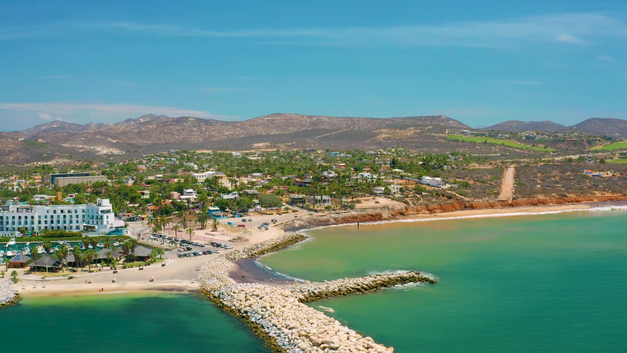 Aerial San Jose Del Cabo shoreline view with the white stone breakwater angled across the lower frame, pale beach and resort buildings at left, green water at right, low development inland, and no visible people.