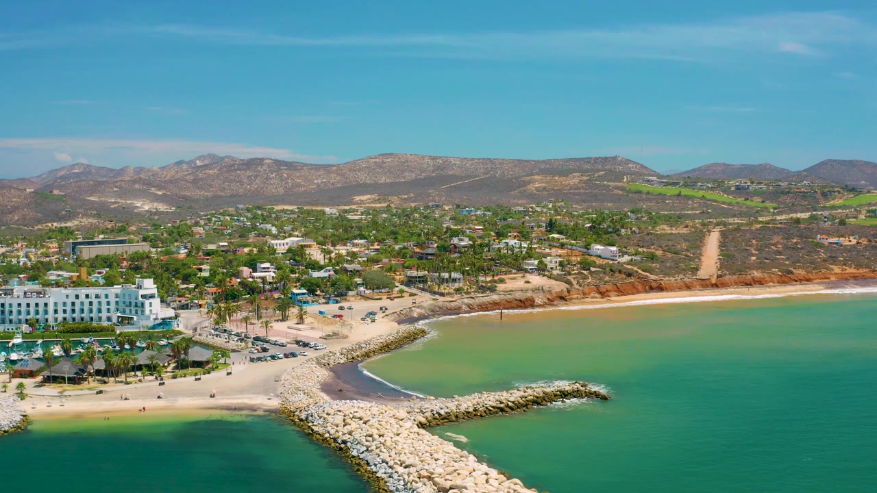 San Jose Del Cabo aerial frame with calm green water surrounding a white rock breakwater, a sandy beach and resort complex at left, palms near the shoreline, hills in the distance, and no visible people.