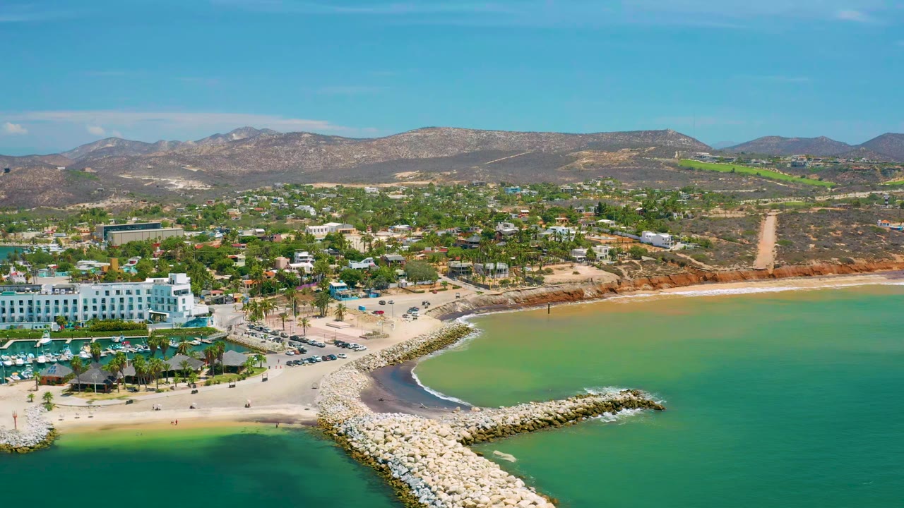 Drone view of beach cove, breakwater rocks, white hotel-like buildings, and hills.