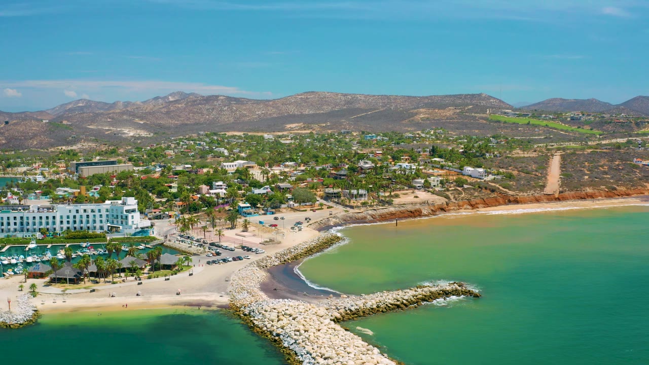 San Jose Del Cabo aerial view with the breakwater lower center, a green lagoon-like patch of shoreline water, pale beach, resort-scale buildings, town streets, hills, and no visible people.