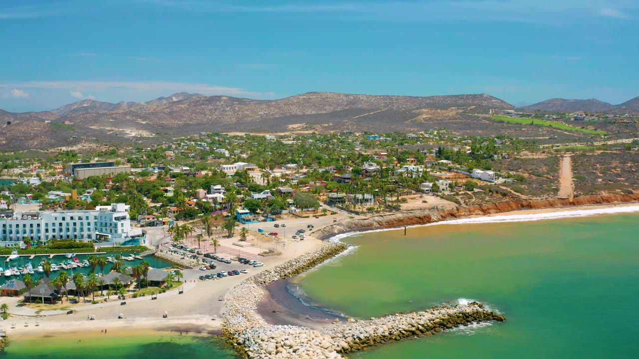 Wide aerial view of a hotel, beach curve, rock jetty, town, and hills.