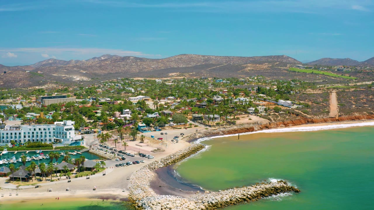 San Jose Del Cabo aerial frame showing the white resort building near the left edge, a sandy beach and breakwater around muted green water, inland neighborhoods, brown hills, and no visible people.