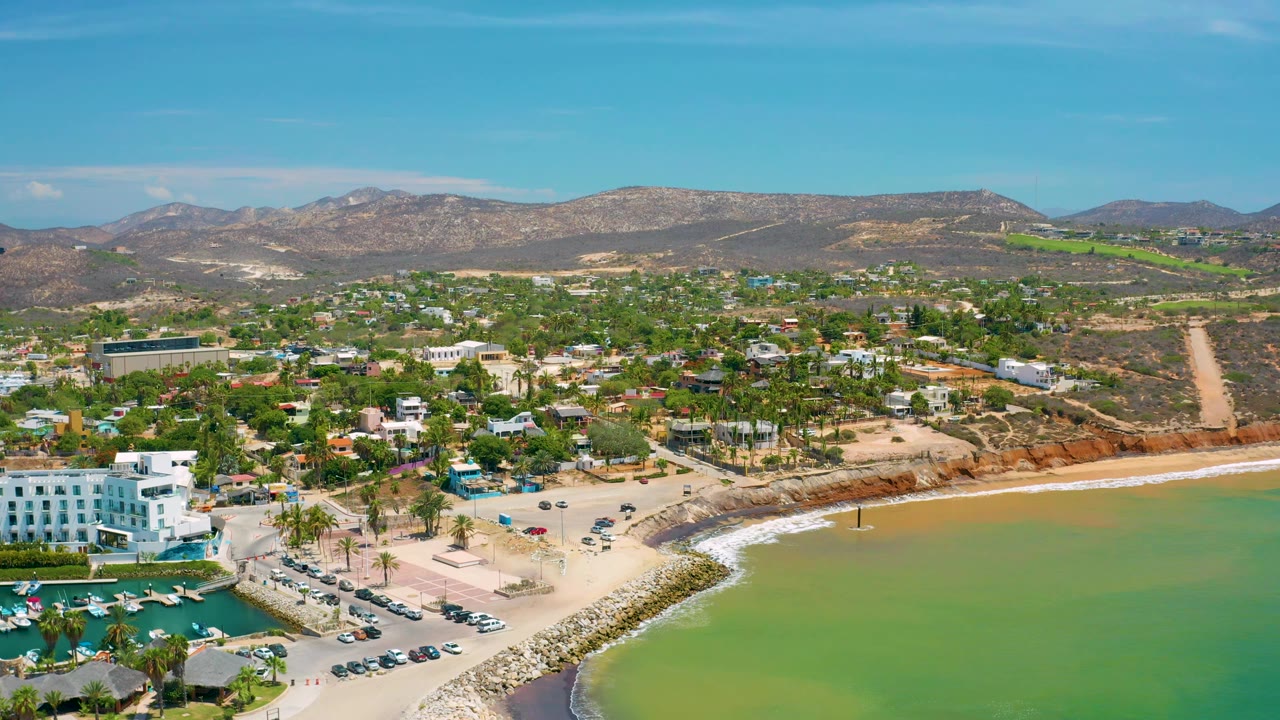 San Jose Del Cabo aerial frame with the beach and breakwater crossing the foreground, green water at right, low buildings and homes in the midground, and no visible people.