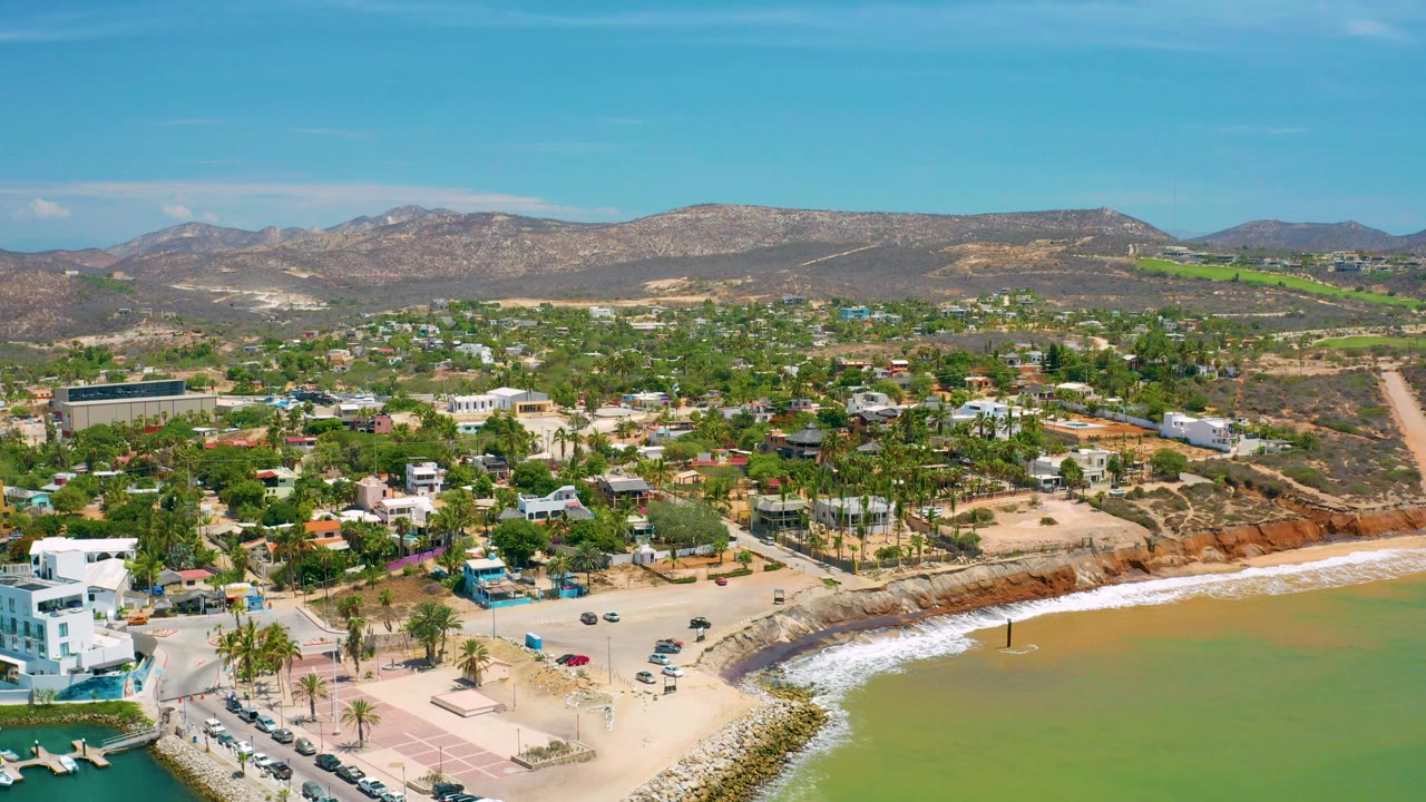Aerial frame over San Jose Del Cabo, Mexico, showing a curved sandy beach, green water, palm-lined streets, low buildings, dry hills, blue sky, and no visible people.