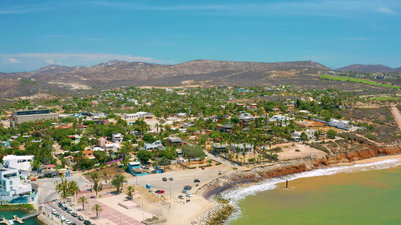 The frame looks across San Jose Del Cabo, Mexico, from the coast toward palm clusters, white and earth-toned buildings, arid hills, green water, and no visible people.