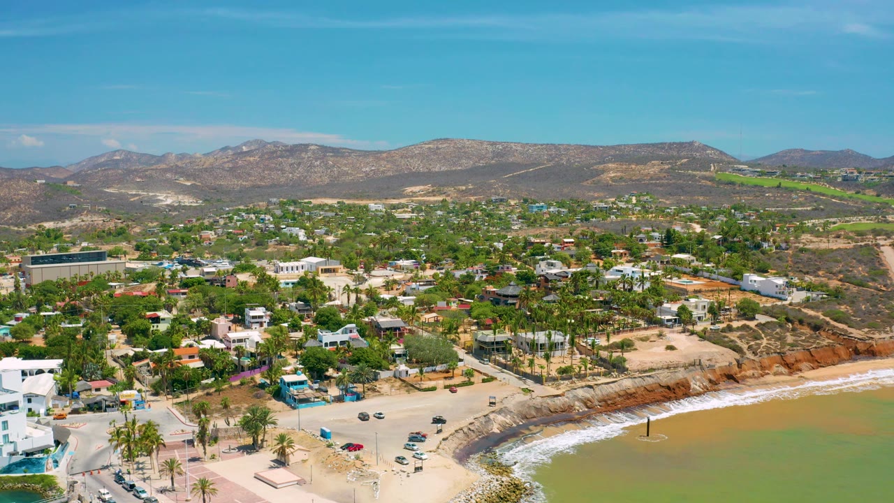 Wide drone view of coastal town, beach curve, palms, and mountain hills.