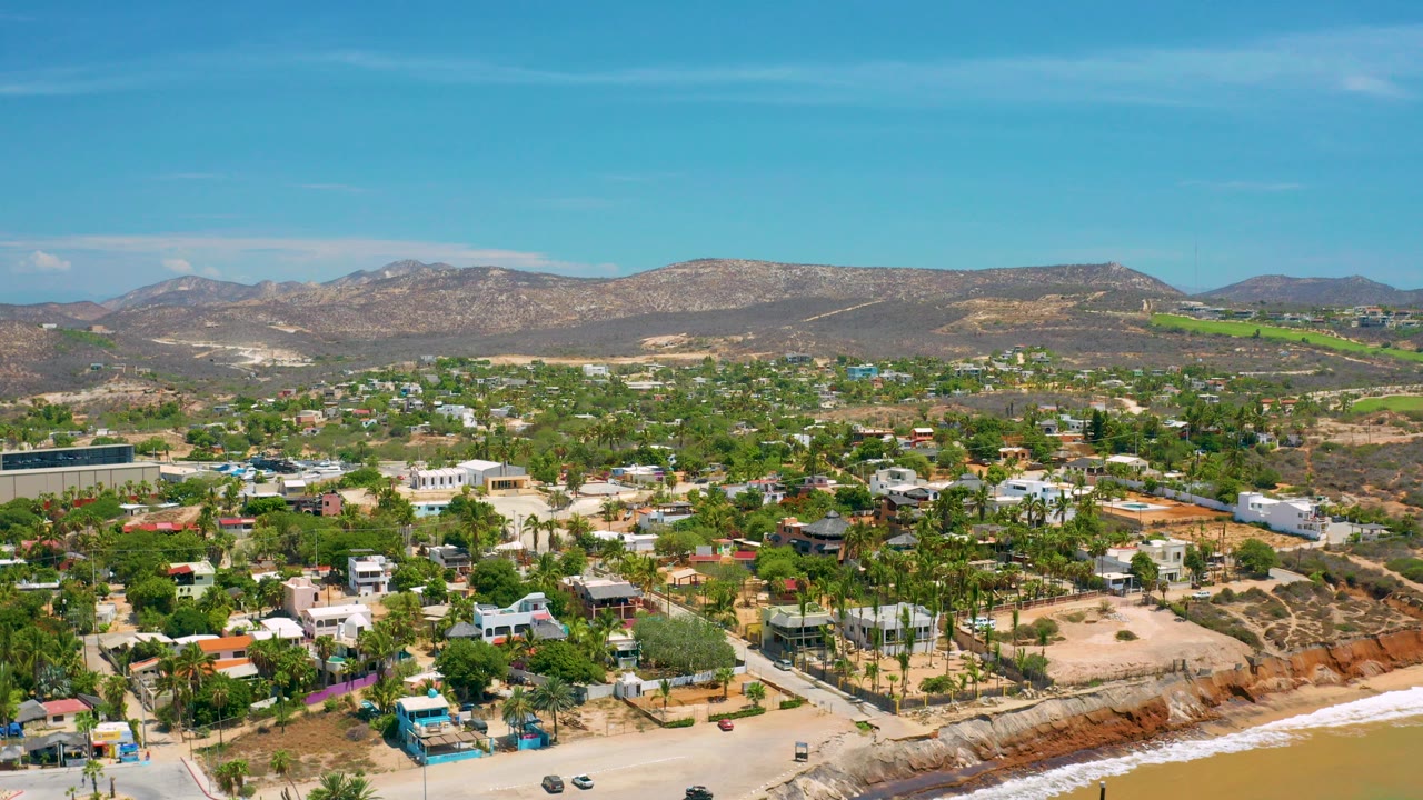 Aerial frame over San Jose Del Cabo showing low-rise coastal homes, palms, sandy lots, a narrow surf line, dry hills, blue sky, and no visible people.