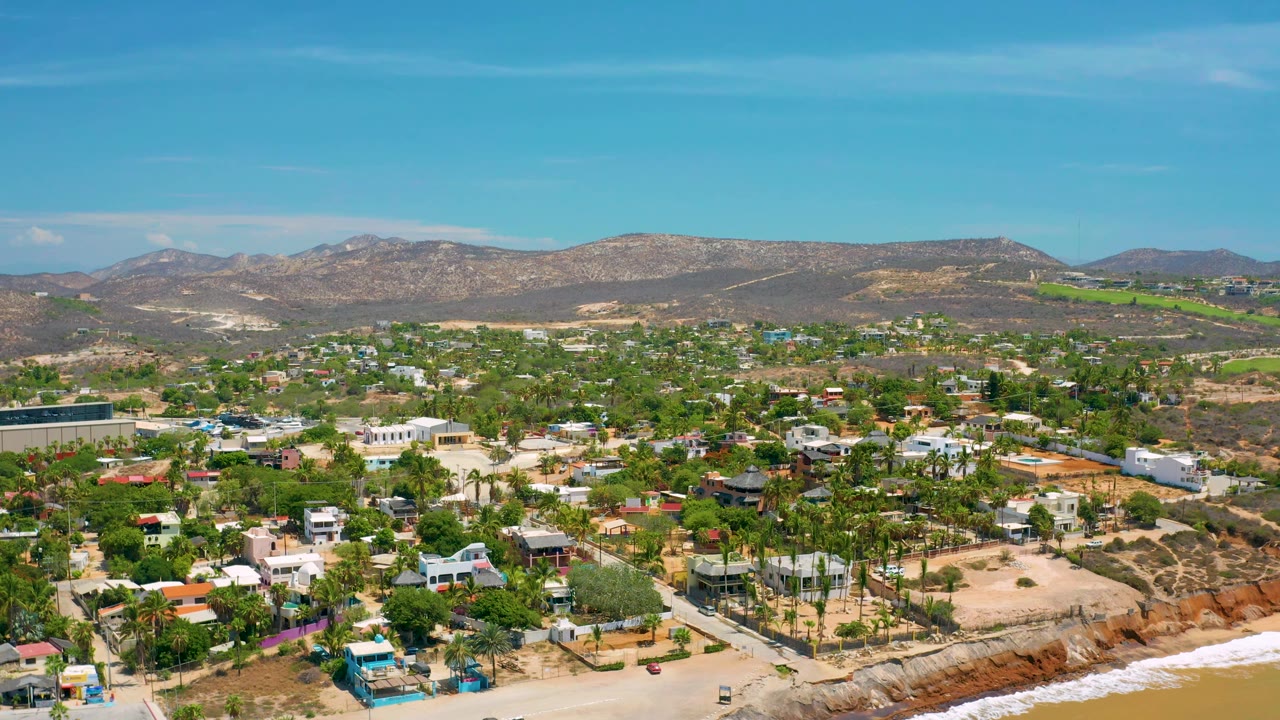Wide San Jose Del Cabo aerial showing ocean-front property, scattered homes, green vegetation, brown hills, clear sky, and no visible people in the frame.