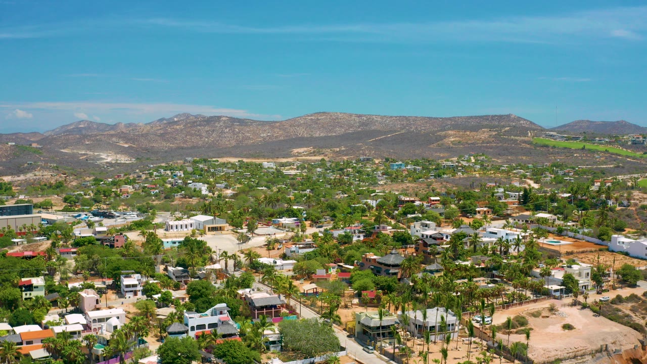Opening aerial frame over San Jose Del Cabo showing low buildings, palm trees, dry lots, distant mountains, and no visible people.
