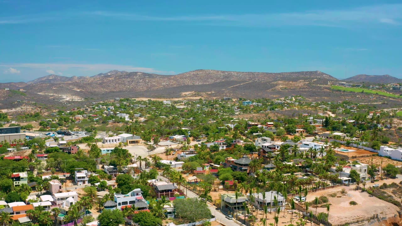 Aerial frame in San Jose Del Cabo with more of the neighborhood grid visible, green palms across the midground, brown hills behind, and no visible people.