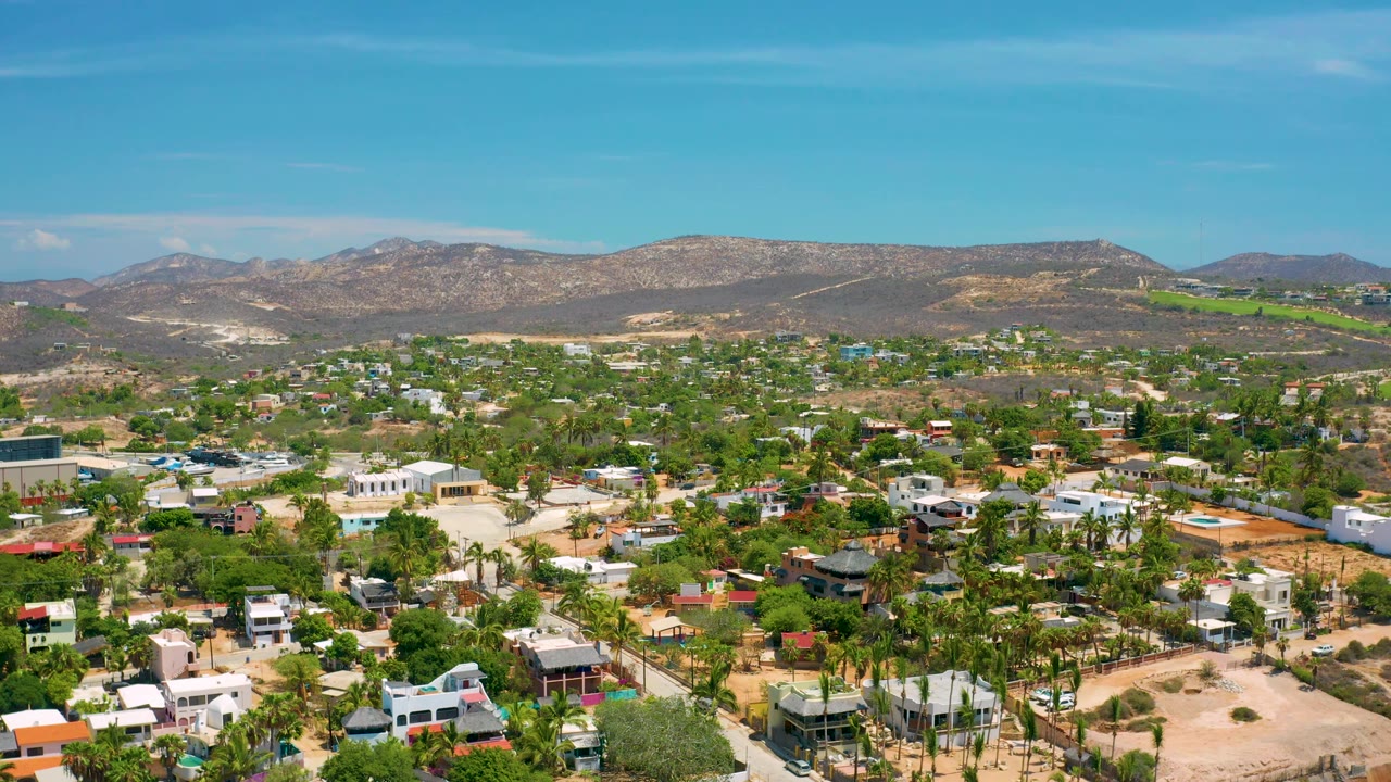 San Jose Del Cabo aerial view with white and colored rooftops in the foreground, dense palms through the center, a mountain ridge on the horizon, and no visible people.