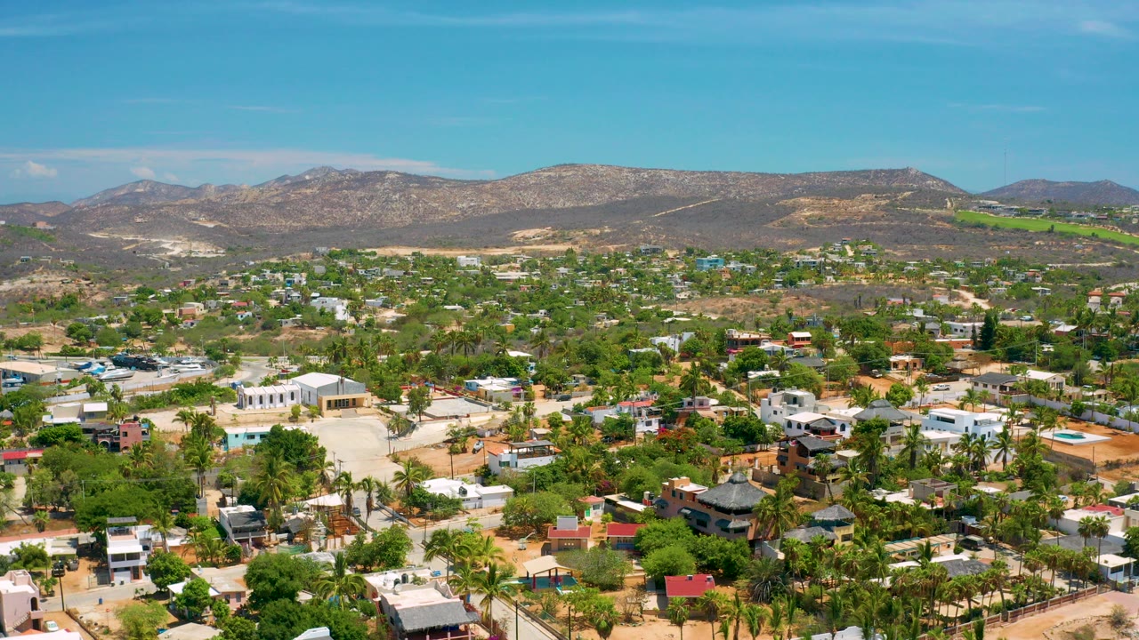 Opening aerial frame over San Jose del Cabo, Mexico, with palms, low-rise homes, sandy streets, distant brown mountains, blue sky, and no visible people.