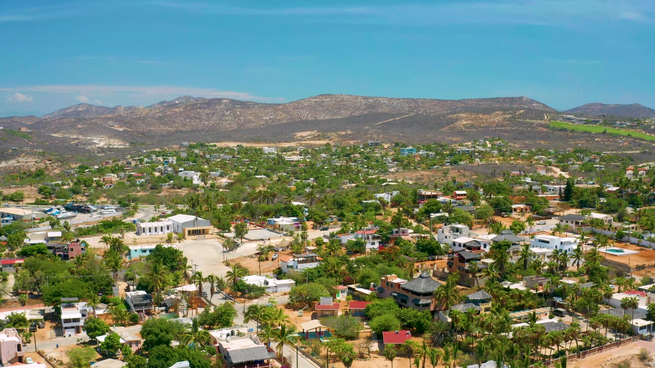 Early aerial frame of San Jose del Cabo showing dense green tree cover, white and pastel buildings, dry mountain ridges, blue sky, and no visible people.