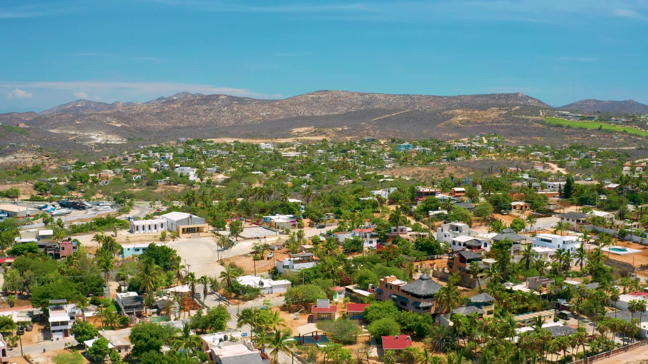 Mid-early aerial frame over San Jose del Cabo with scattered pools, palms, flat-roofed buildings, dry roads, brown mountains, blue sky, and no visible people.