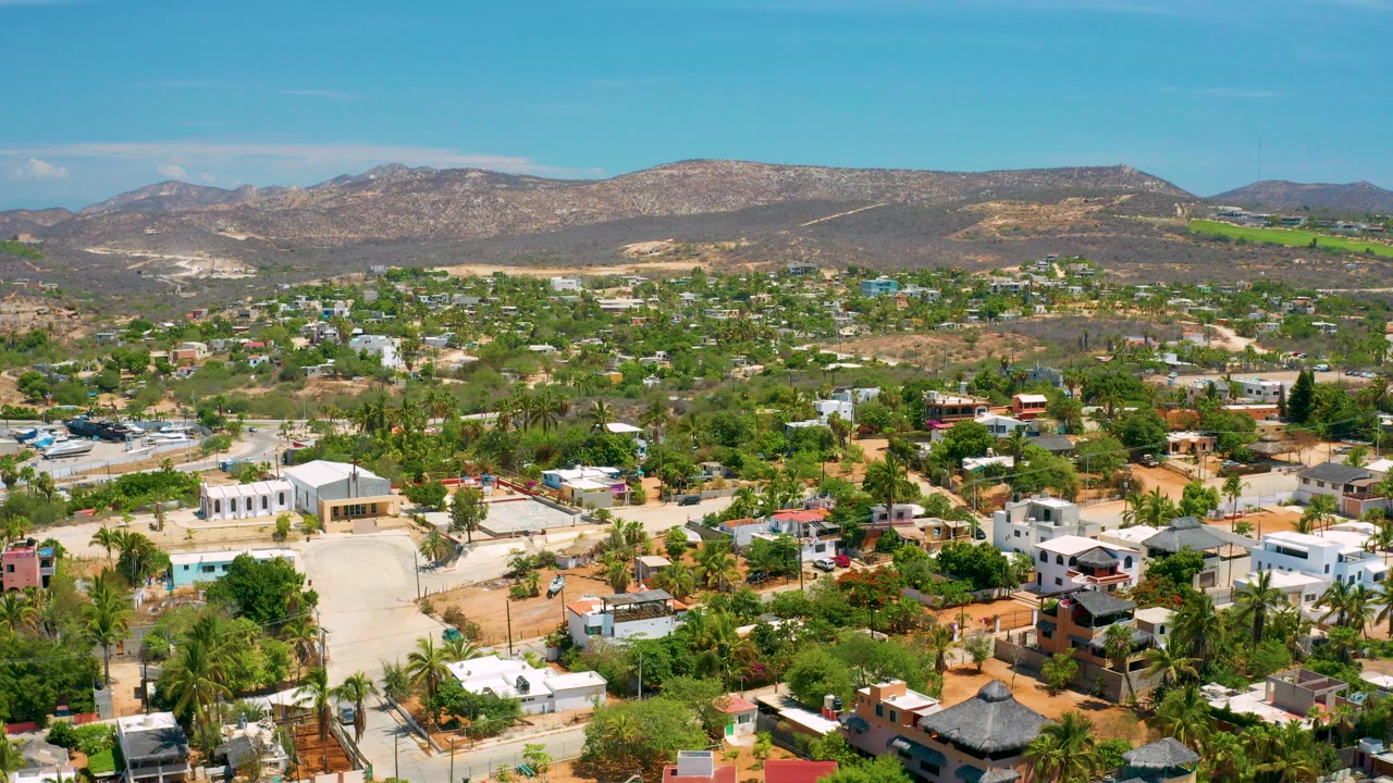 Opening aerial frame over San Jose Del Cabo showing low-rise buildings mixed with trees and palms, dry tan ground, distant hills, blue sky, and no visible people.