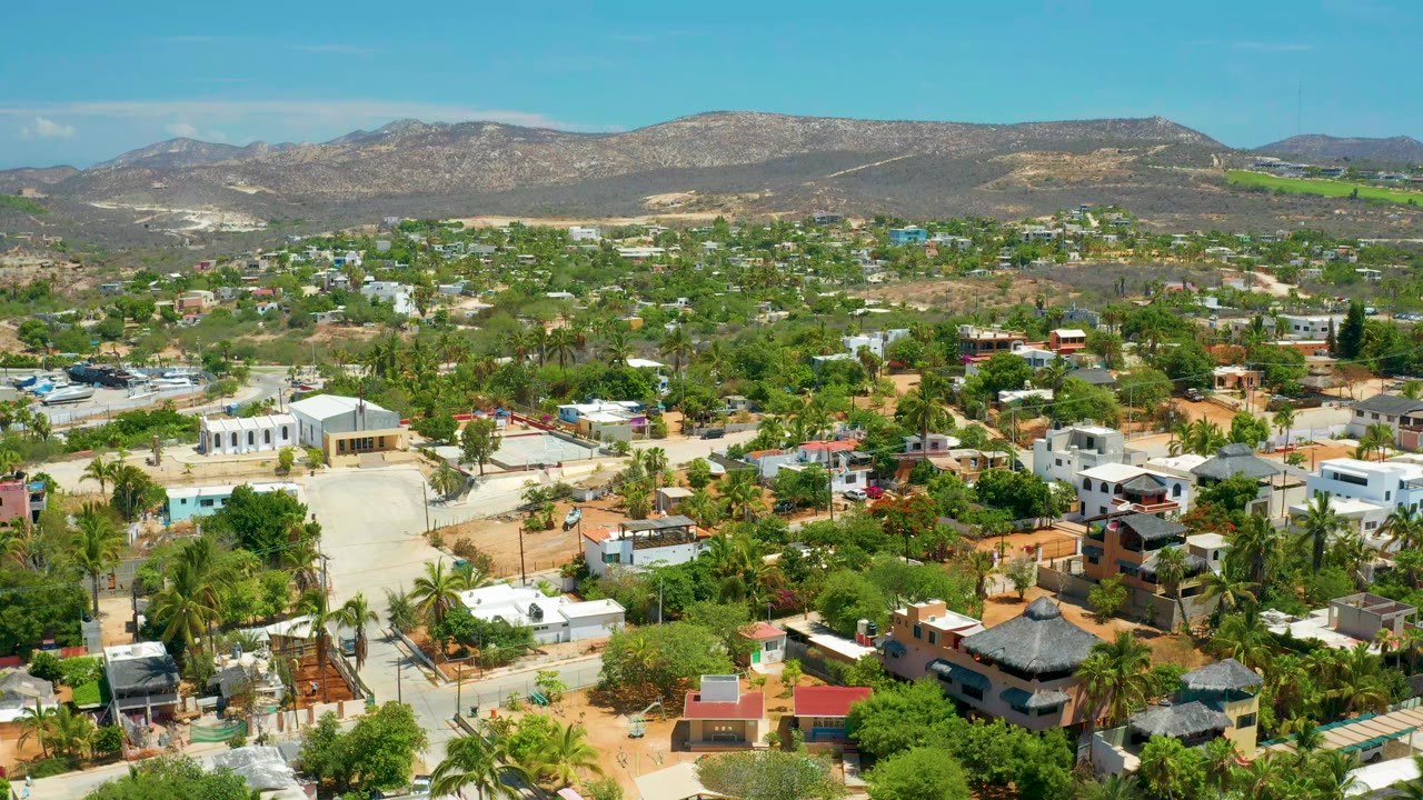 Aerial view over San Jose Del Cabo, Mexico, showing palms, low-rise homes, dirt roads, dry hills, blue sky, and no visible people at the opening moment.