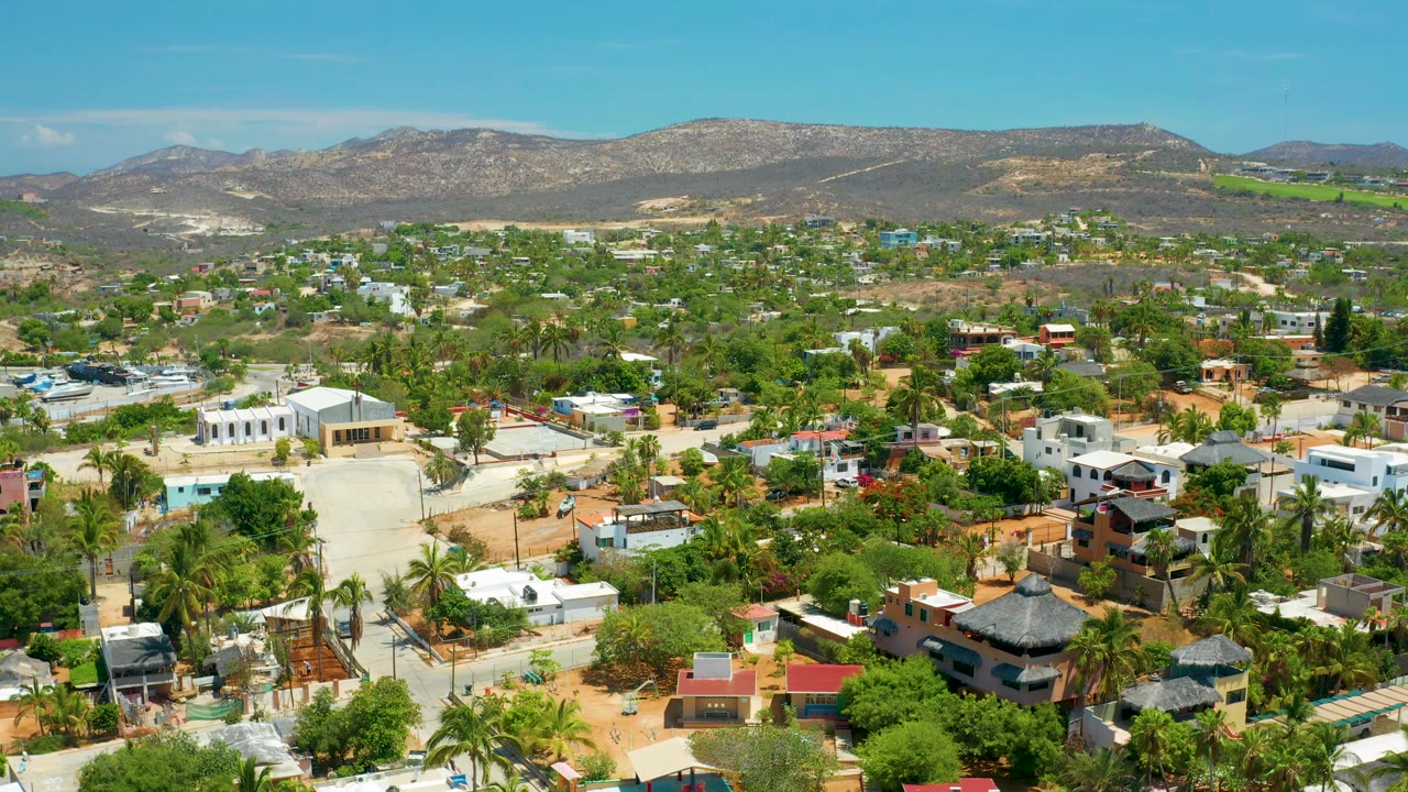 Elevated San Jose Del Cabo view with clustered houses, palms, sandy roads, green vegetation, a long dry ridge, and no visible people.