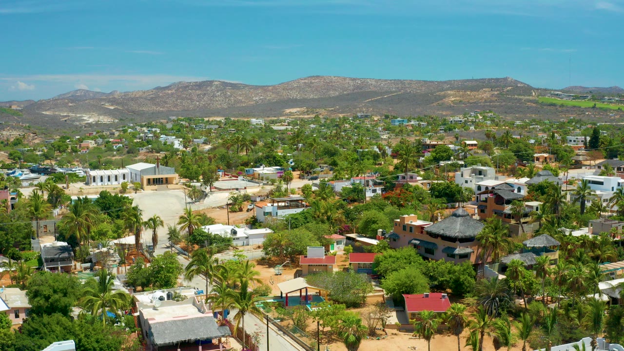 Aerial view over San Jose Del Cabo homes and palm-lined streets, with dry hills across the horizon and no visible people.