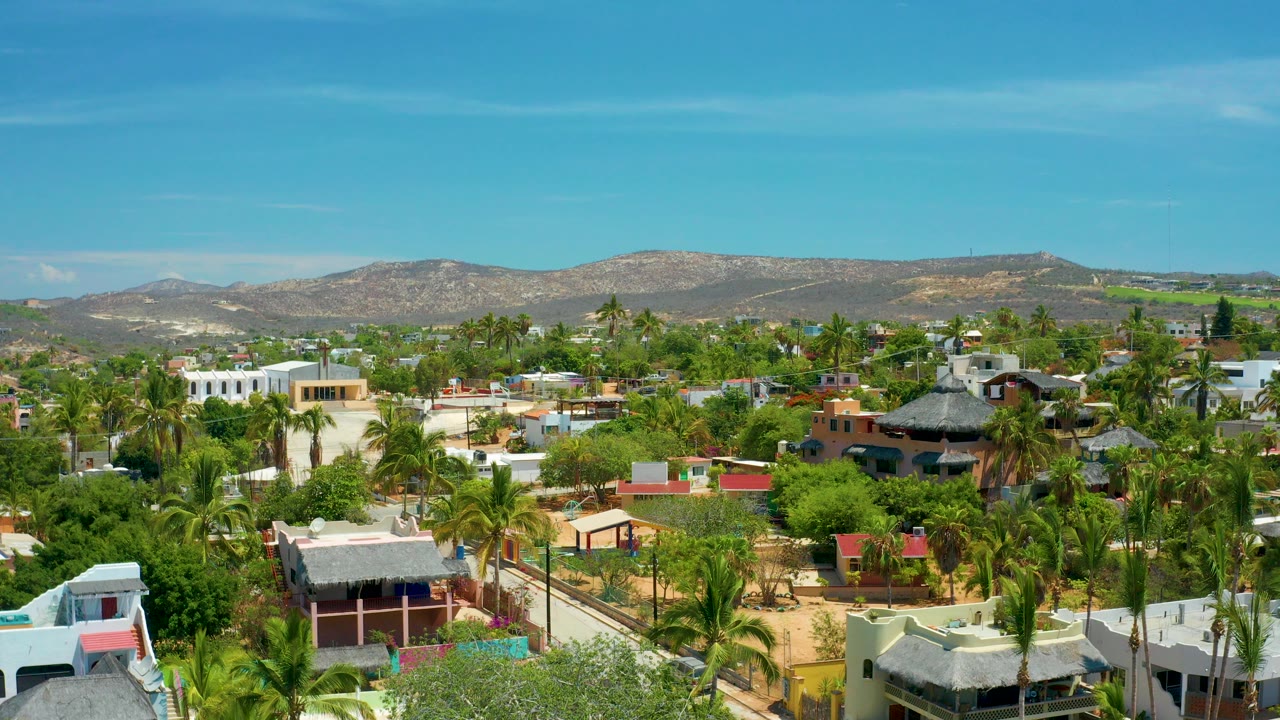 Opening elevated view over San Jose Del Cabo with palms, low homes, thatched roofs, dry hills, blue sky, and no visible people.