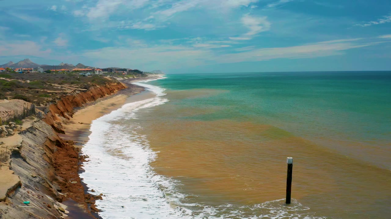 San Jose del Cabo shore with foamy waves pushing farther up the beach, layered reddish cliffs on the left, shallow green-blue water across the frame, the black post near the lower right, and no visible people.